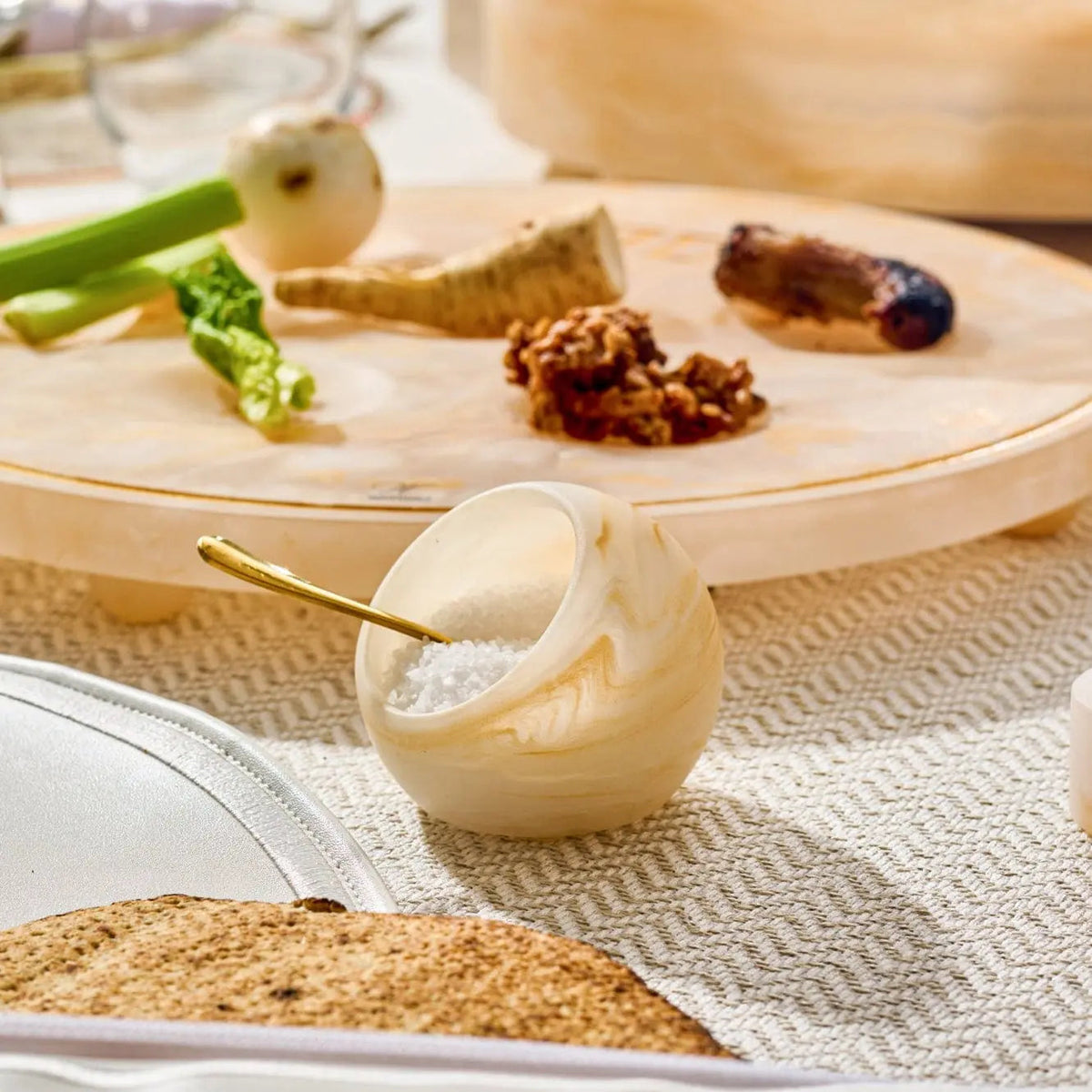 Cream resin salt cellar with gold spoon sitting on neutral linens, surrounded by fresh herbs and spices on wooden board.