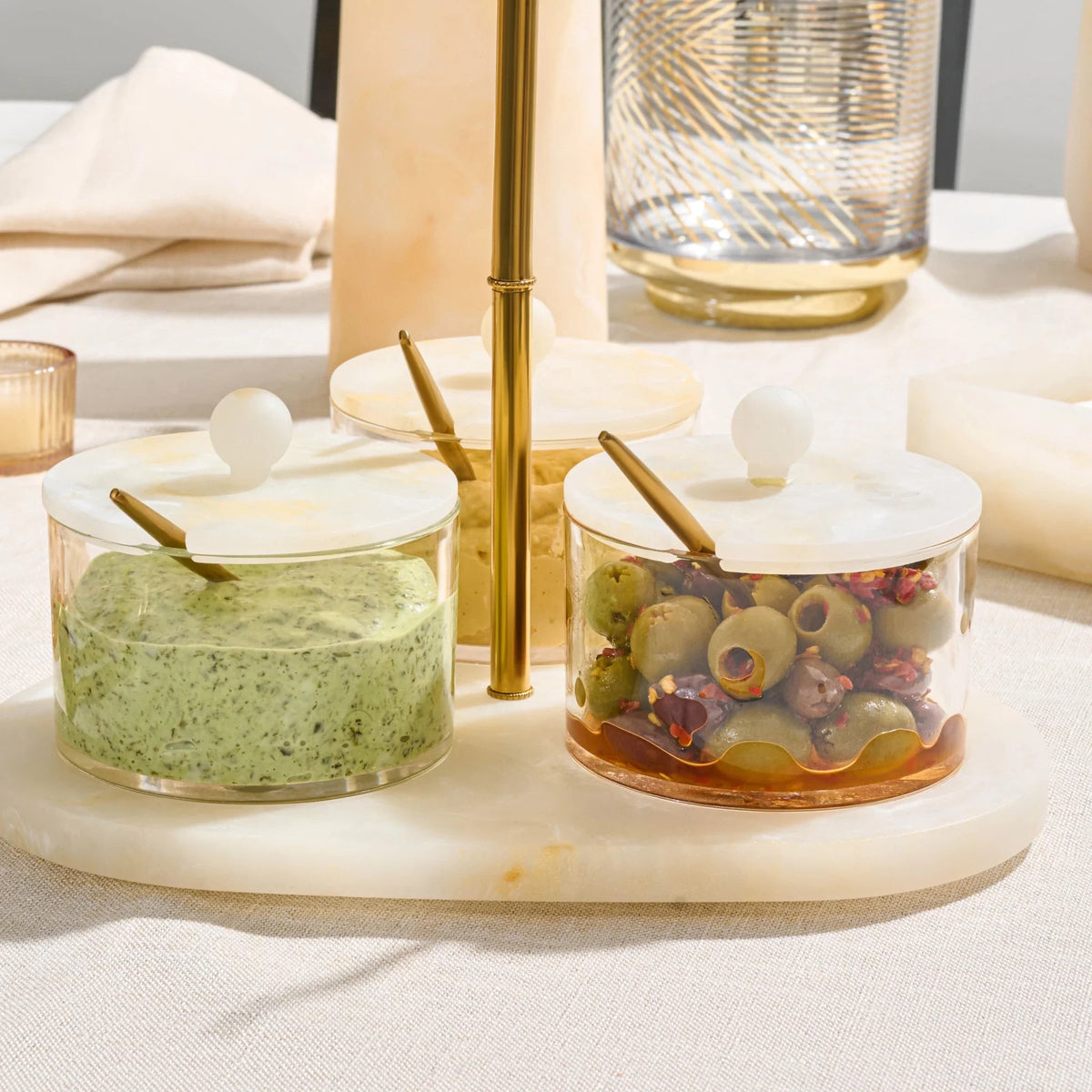 Three clear glass condiment jars with resin lids and gold spoons, arranged on a round roesin tray. The jars contain green herb spread, a yellow spread, and sliced olives with red peppers, set on a beige tablecloth with dining props in the background.