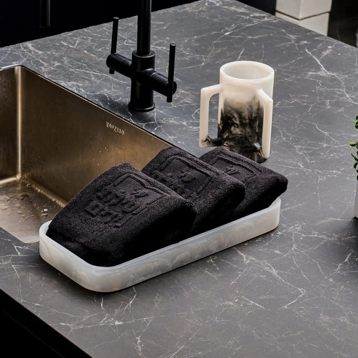 Cream and black resin towel tray with folded dark towels, displayed on a marble bathroom countertop with toiletries nearby.