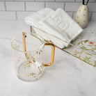 Floral Bird Washing Cup in clear glass with gold handles, displayed with white towels and home decor on marble surface