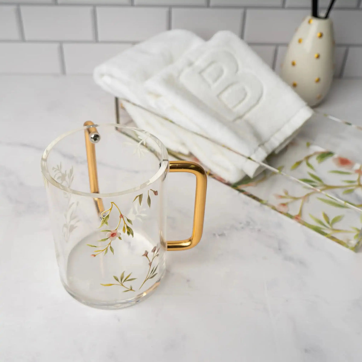 Floral Bird Washing Cup in clear glass with gold handles, displayed with white towels and home decor on marble surface