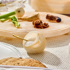 Cream resin salt cellar with gold spoon sitting on neutral linens, surrounded by fresh herbs and spices on wooden board.