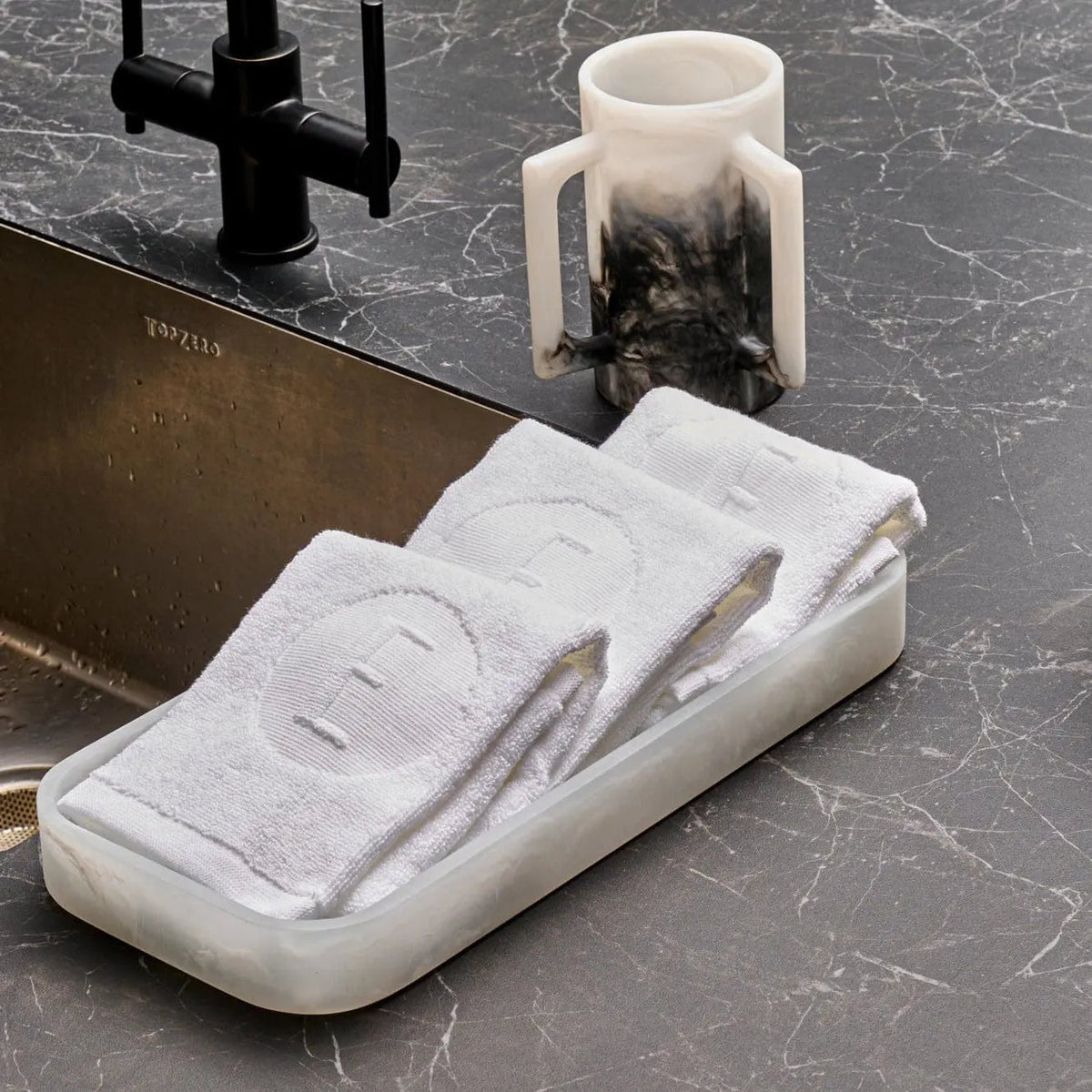 White embossed circle initial finger towel folded on marble counter with bathroom accessories nearby