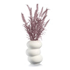 White ceramic donut vase with stacked ring design holding dried purple faux flowers and grasses arrangement.