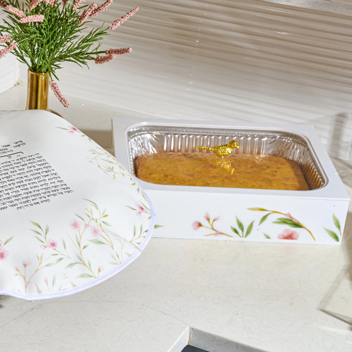  light countertop, with  a rectangular foil pan in a white acrylic  floralpan holder  holding a golden bird with a cake inside