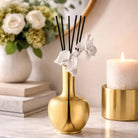 Gold pear-shaped Aura Orchid Vase Diffuser with black reeds and white orchid flowers, styled with candles and books on a white surface.