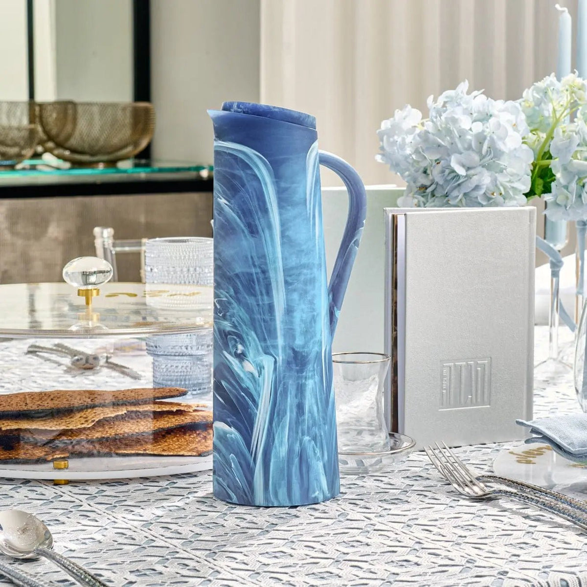 Blue resin pitcher with marbled swirl pattern and textured handle, displayed on a table setting with tabletop decor by Waterdale Collection.