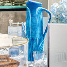 Blue resin pitcher with handle from Waterdale Collection, displayed on a white tabletop with decorative home accessories.