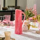 Coral pink resin pitcher with textured surface displayed on a marble table setting with glassware and floral centerpiece.