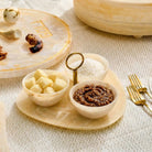 Beige resin dip bowl set with two small bowls holding nuts and chocolate pieces, displayed on natural tabletop with serving utensils and wooden vessels nearby.