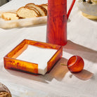 A set of amber resin  tableware on a white tablecloth, including a square napkin holder with white napkins, a tall resin pitcher , and a small salt cellar 