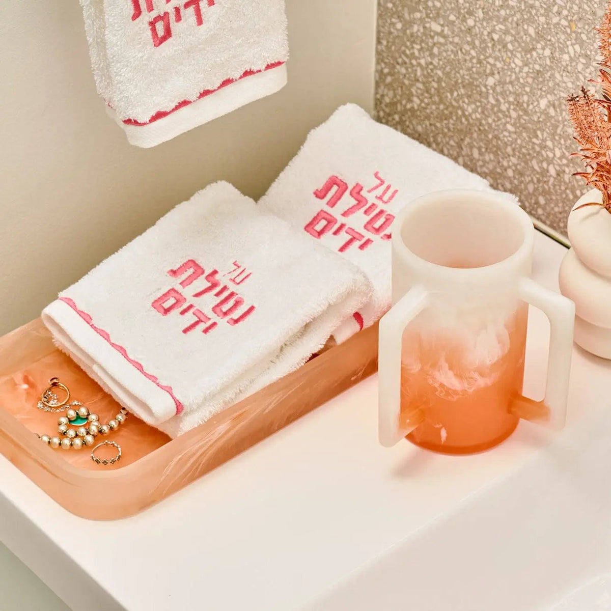 Cream resin tray with pink Hebrew text displayed on bathroom vanity next to towels and a coral drink