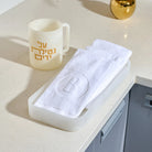 A white textured towel with an embroidered letter 'B' inside a circle, folded in a translucent white resin tray , set on a light beige countertop next to a cream-colored resin washing cup  with gold Hebrew text.