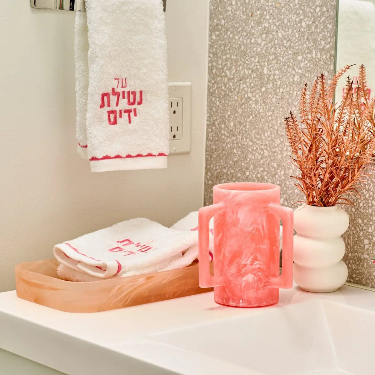 Pink resin washing cup displayed on a wooden tray with white towels and dried florals in a bathroom vanity setting.