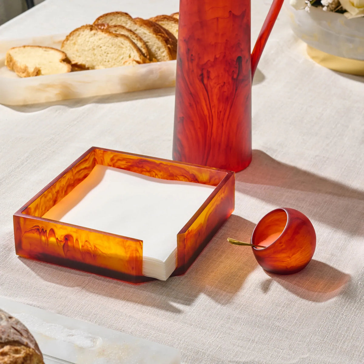 A set of amber marbled glass tableware on a white tablecloth, including a square napkin holder with white napkins, a resin amber pitcher  , and a small resin amber salt cellar