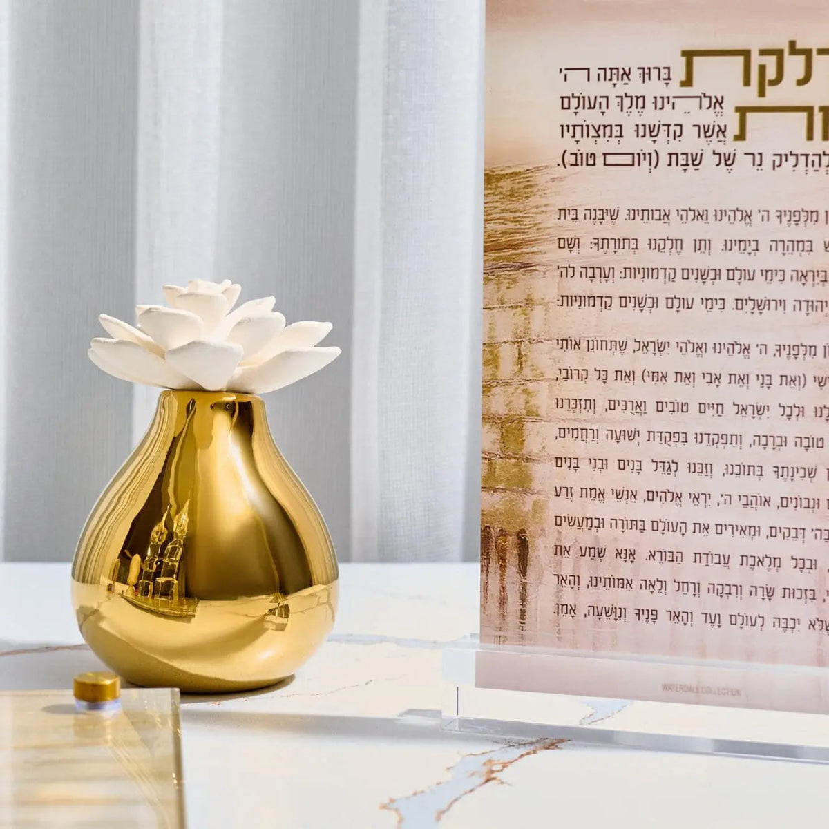 Gold teardrop-shaped flower diffuser with white cap displayed beside Hebrew prayer book, perfect for home fragrance and housewarming gifts.