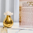 Gold teardrop-shaped flower diffuser with white cap displayed beside Hebrew prayer book, perfect for home fragrance and housewarming gifts.