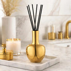 Gold Aura Vase Diffuser with black reed sticks on marble surface, surrounded by candles and home decor accents.