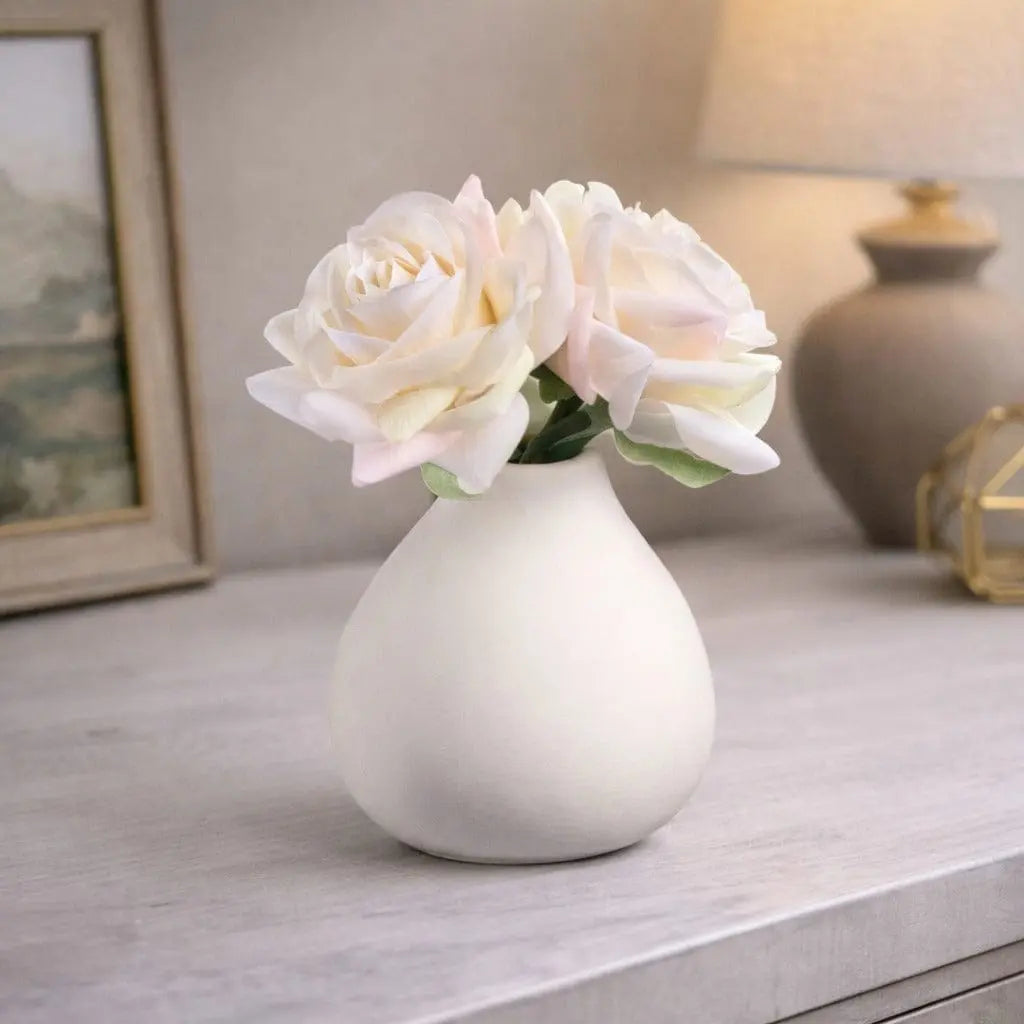 White ceramic teardrop vase with faux cream roses arranged on a marble surface, styled with home décor accessories in the background.