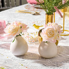 White faux floral teardrop vase with cream and blush roses arranged on a styled tabletop setting with gold accents.