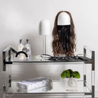 Lucite wig head display stand holding long wavy dark hair wig, shown on clear acrylic shelf unit with storage below, styled with beauty accessories and plants.